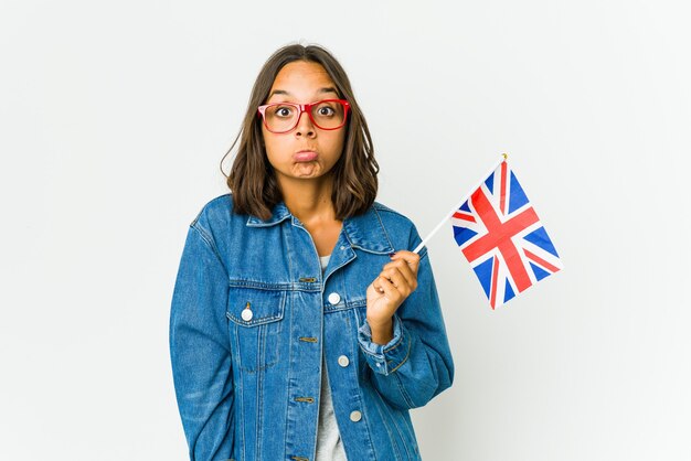 Mujer joven sosteniendo una bandera inglesa aislada en la pared blanca se encoge de hombros y abre los ojos confundidos