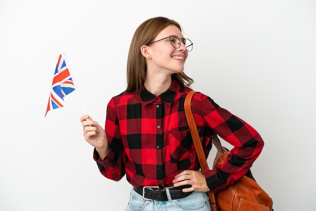 Mujer joven sosteniendo una bandera del Reino Unido aislada de fondo azul posando con los brazos en la cadera y sonriendo