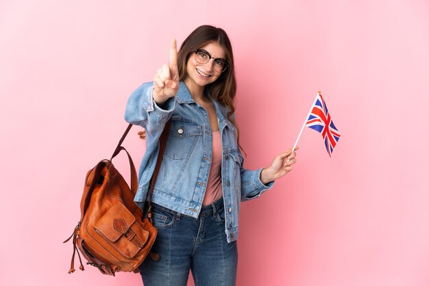 Mujer joven sosteniendo una bandera del Reino Unido aislada en rosa mostrando y levantando un dedo