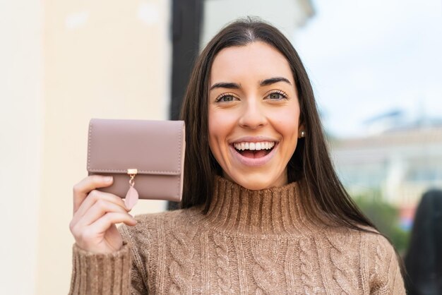 Mujer joven sosteniendo una billetera al aire libre con sorpresa y expresión facial conmocionada