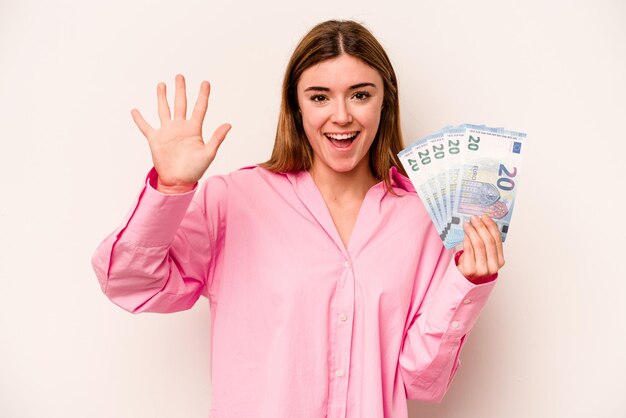 Mujer joven sosteniendo billetes aislados sobre fondo blanco sonriendo alegre mostrando el número cinco con los dedos