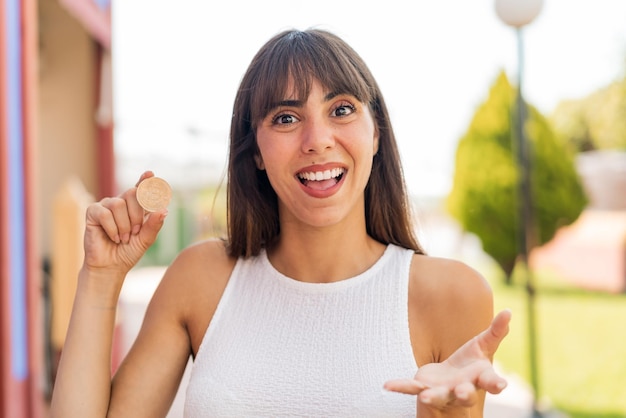 Mujer joven sosteniendo un Bitcoin al aire libre con expresión facial sorprendida