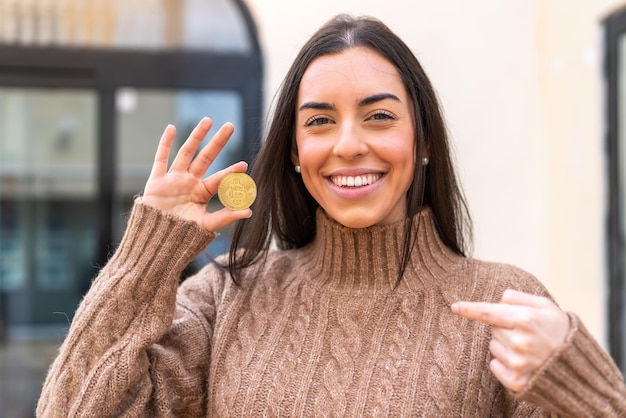 Mujer joven sosteniendo un Bitcoin al aire libre con expresión facial sorpresa