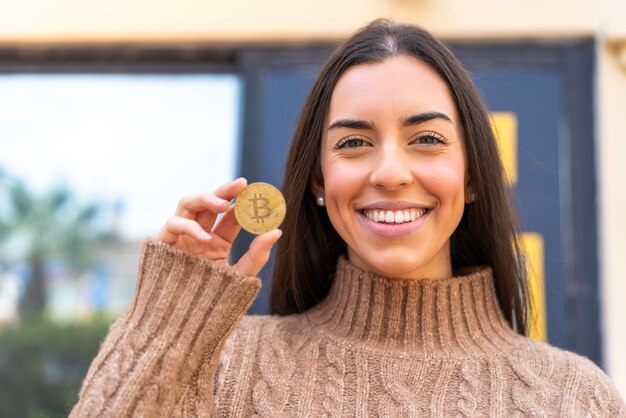 Mujer joven sosteniendo un Bitcoin al aire libre sonriendo mucho