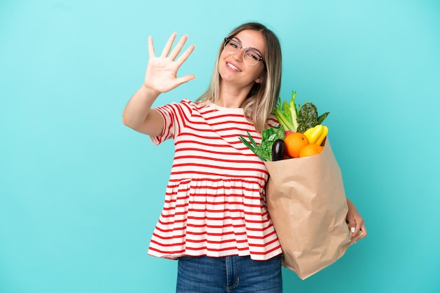 Mujer joven sosteniendo una bolsa de compras aislada de fondo azul contando cinco con los dedos