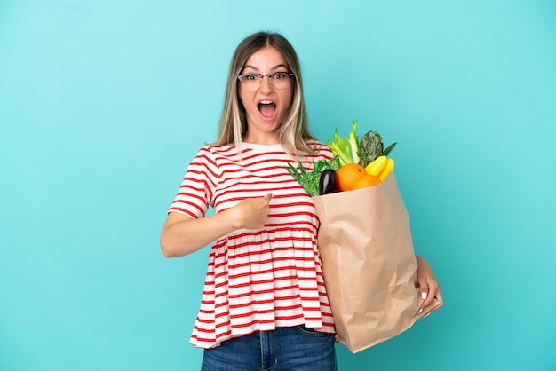 Mujer joven sosteniendo una bolsa de compras aislada de fondo azul con expresión facial sorpresa