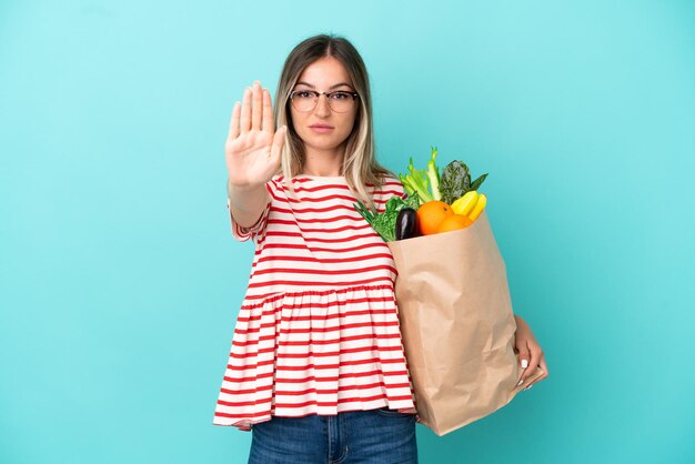 Mujer joven sosteniendo una bolsa de compras aislada de fondo azul haciendo un gesto de parada