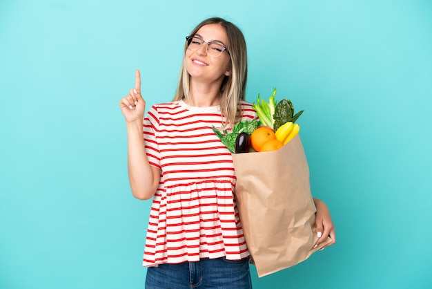 Mujer joven sosteniendo una bolsa de compras aislada en un fondo azul mostrando y levantando un dedo en señal de lo mejor