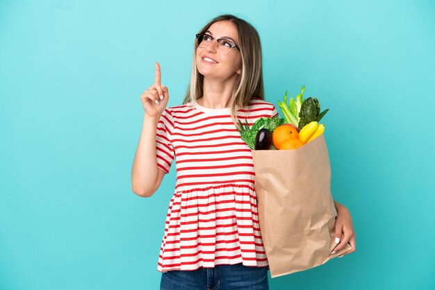 Mujer joven sosteniendo una bolsa de compras aislada de fondo azul señalando una gran idea