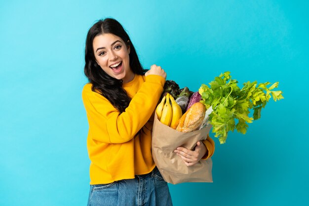 Mujer joven sosteniendo una bolsa de compras aislada en la pared azul celebrando una victoria