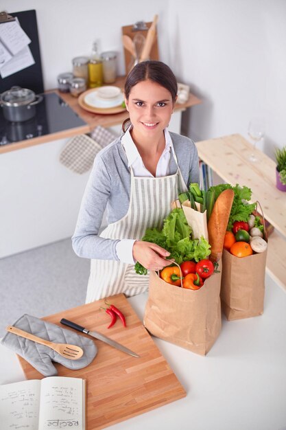 Mujer joven sosteniendo la bolsa de compras con verduras de pie en la cocina