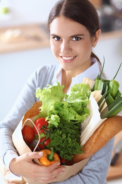 Mujer joven sosteniendo la bolsa de compras con verduras de pie en la cocina