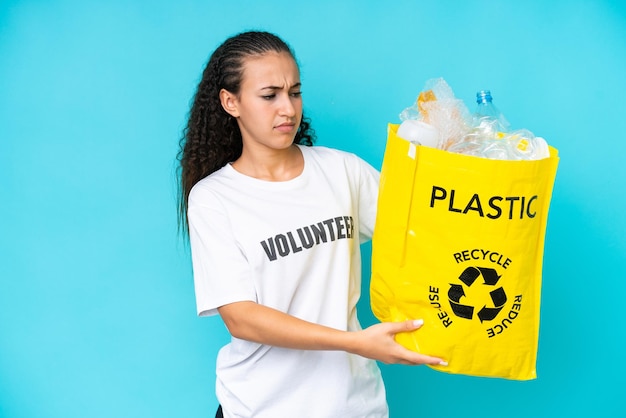 Mujer joven sosteniendo una bolsa llena de botellas de plástico para reciclar aislado sobre fondo azul con expresión triste