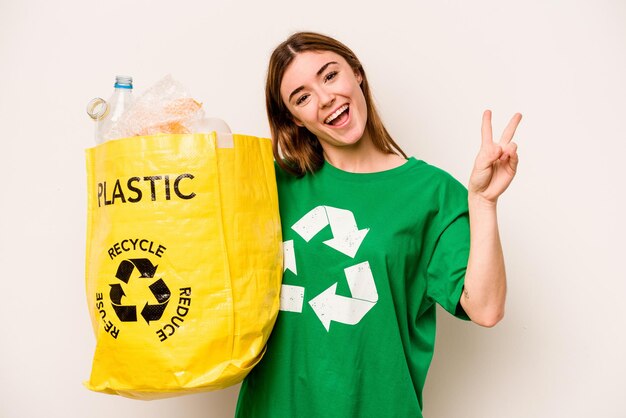 Mujer joven sosteniendo una bolsa llena de botellas de plástico para reciclar aislado sobre fondo blanco alegre y despreocupado mostrando un símbolo de paz con los dedos