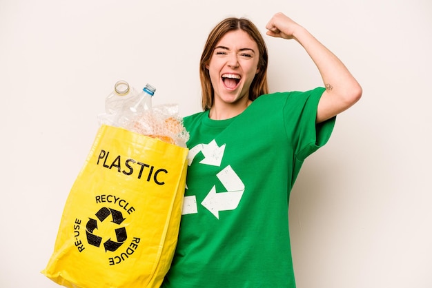 Mujer joven sosteniendo una bolsa llena de botellas de plástico para reciclar aislado sobre fondo blanco levantando el puño después de un concepto de ganador de la victoria