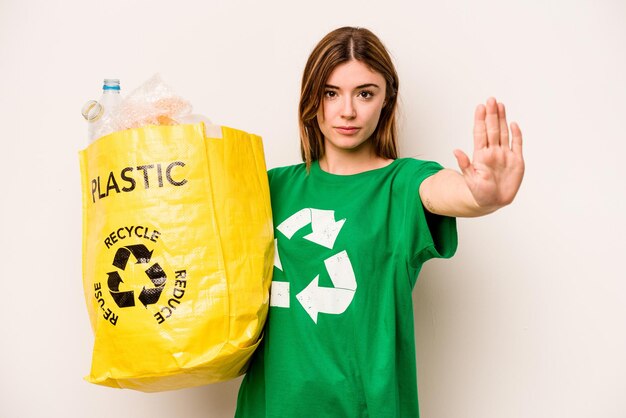 Foto mujer joven sosteniendo una bolsa llena de botellas de plástico para reciclar aislado sobre fondo blanco de pie con la mano extendida mostrando la señal de stop que le impide