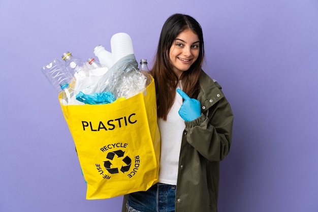 Mujer joven sosteniendo una bolsa llena de botellas de plástico para reciclar aislado sobre fondo púrpura con expresión facial sorpresa