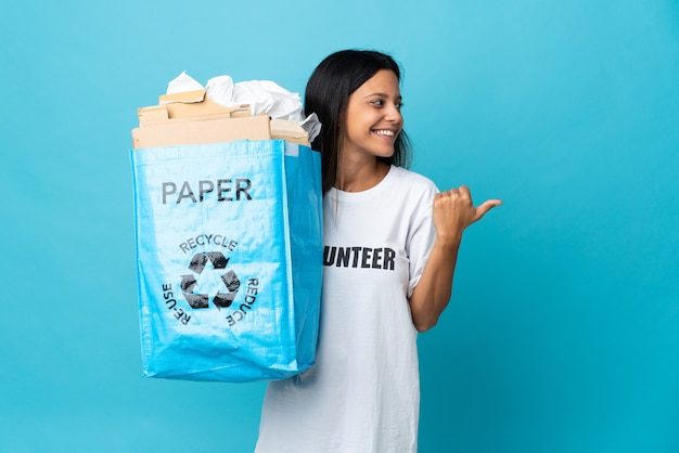 Mujer joven sosteniendo una bolsa de reciclaje llena de papel apuntando hacia el lado para presentar un producto