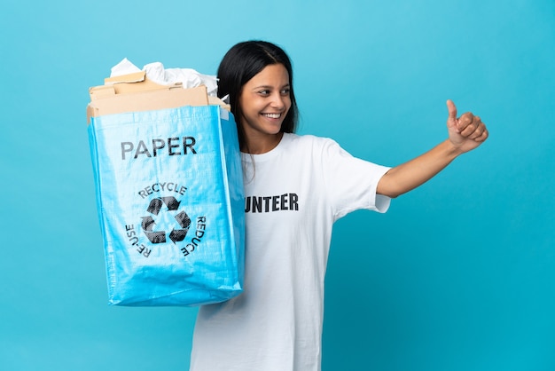 Mujer joven sosteniendo una bolsa de reciclaje llena de papel dando un pulgar hacia arriba gesto