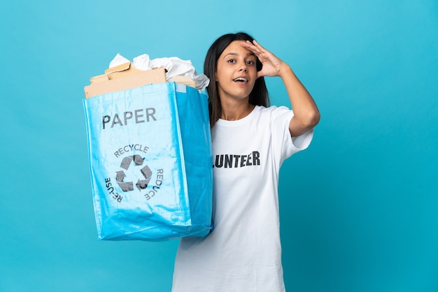 Mujer joven sosteniendo una bolsa de reciclaje llena de papel haciendo gesto de sorpresa mientras mira hacia un lado