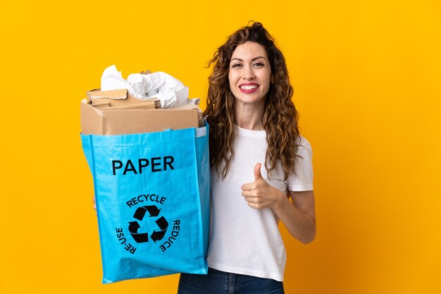 Mujer joven sosteniendo una bolsa de reciclaje llena de papel para reciclar aislado en amarillo dando un pulgar hacia arriba gesto