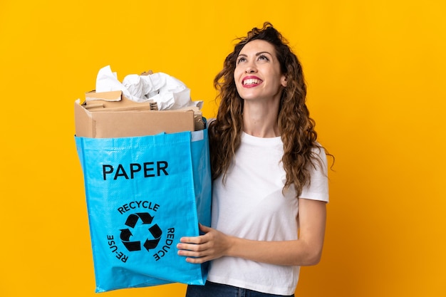 Mujer joven sosteniendo una bolsa de reciclaje llena de papel para reciclar aislado en amarillo pensando en una idea mientras mira hacia arriba