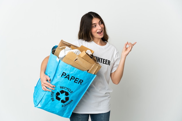 Mujer joven sosteniendo una bolsa de reciclaje llena de papel para reciclar aislado en blanco con la intención de darse cuenta de la solución mientras levanta un dedo hacia arriba