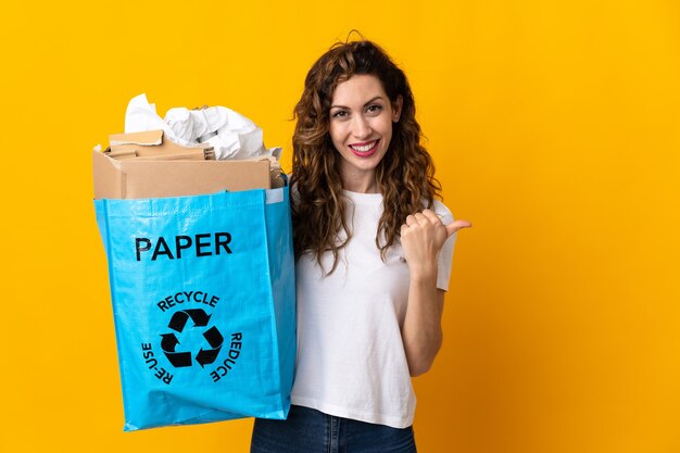 Mujer joven sosteniendo una bolsa de reciclaje llena de papel para reciclar aislado en la pared amarilla apuntando hacia el lado para presentar un producto