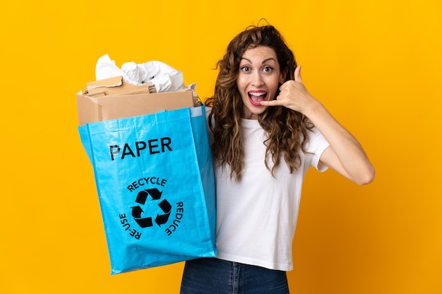 Mujer joven sosteniendo una bolsa de reciclaje llena de papel para reciclar aislado en la pared amarilla haciendo gesto de teléfono.