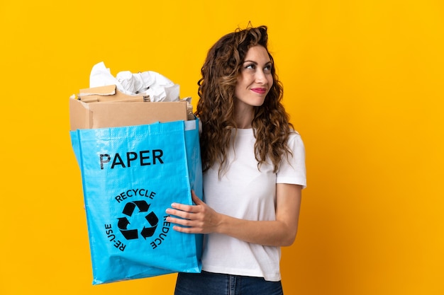 Mujer joven sosteniendo una bolsa de reciclaje llena de papel para reciclar aislado en la pared amarilla mirando hacia arriba mientras sonríe