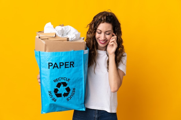 Mujer joven sosteniendo una bolsa de reciclaje llena de papel para reciclar aislado en la pared amarilla riendo