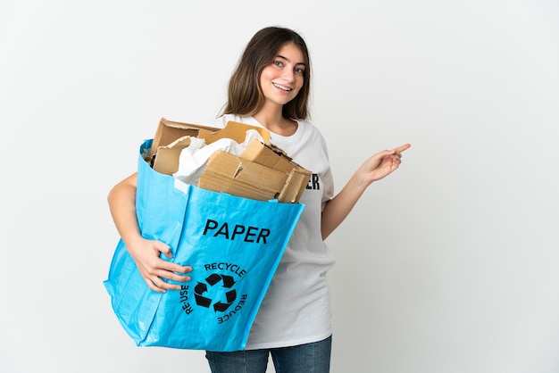 Mujer joven sosteniendo una bolsa de reciclaje llena de papel para reciclar aislado en la pared blanca apuntando con el dedo hacia el lado