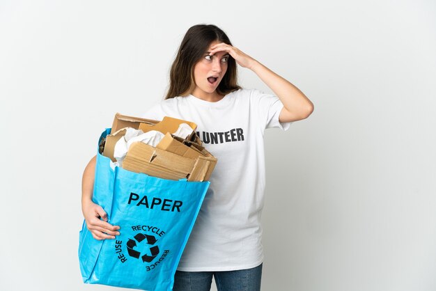 Mujer joven sosteniendo una bolsa de reciclaje llena de papel para reciclar aislado en la pared blanca haciendo un gesto de sorpresa mientras mira hacia el lado