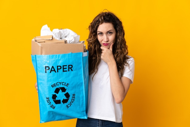 Mujer joven sosteniendo una bolsa de reciclaje llena de papel para reciclar aislado sobre fondo amarillo pensando