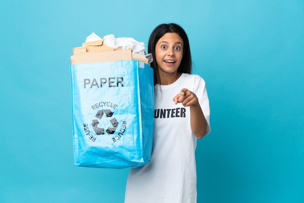 Mujer joven sosteniendo una bolsa de reciclaje llena de papel sorprendido y apuntando hacia el frente