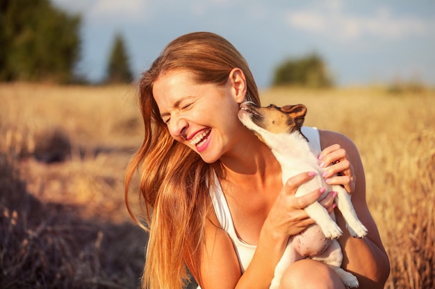 Mujer joven sosteniendo un cachorro Jack Russell terrier en su mano, tratando de posar, pero el perro está lamiendo y masticando su oreja y cabello.