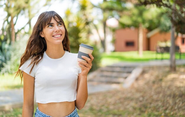 Mujer joven sosteniendo un café para llevar al aire libre mirando hacia arriba mientras sonríe