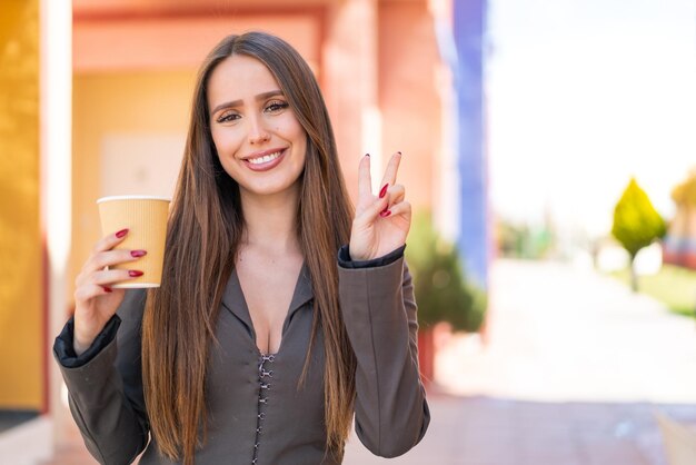 Mujer joven sosteniendo un café para llevar al aire libre sonriendo y mostrando el signo de la victoria