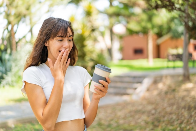 Mujer joven sosteniendo un café para llevar al aire libre con sorpresa y expresión facial conmocionada