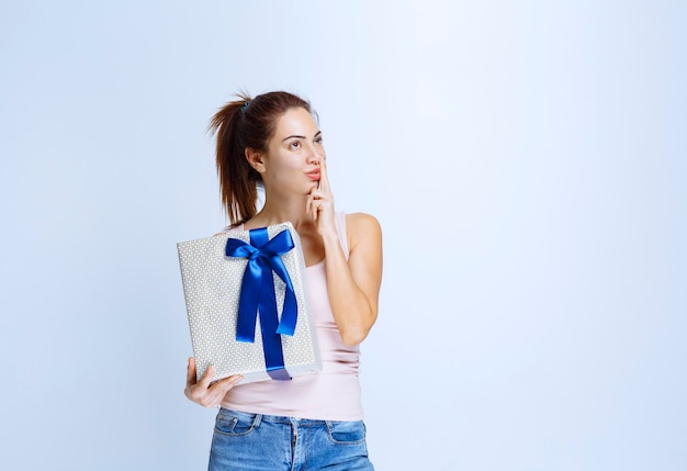 Mujer joven sosteniendo una caja de regalo blanca envuelta con cinta azul y pensando y dudando