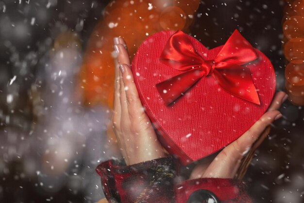 Mujer joven sosteniendo una caja de regalo roja en forma de corazón en la noche durante las nevadas