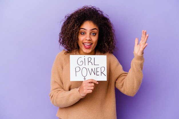 Mujer joven sosteniendo un cartel de Girl power aislado en la pared púrpura recibiendo una agradable sorpresa, emocionado y levantando las manos