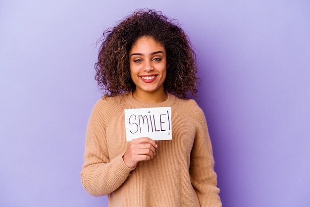 Mujer joven sosteniendo un cartel de sonrisa aislado en la pared púrpura feliz, sonriente y alegre