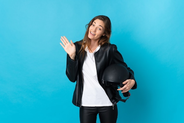 Mujer joven sosteniendo un casco de motocicleta aislado en la pared azul saludando con la mano con expresión feliz