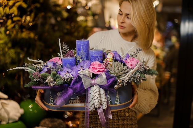 Mujer joven sosteniendo una composición navideña con flores púrpuras y rosas, suculentas, ramas de abeto y velas