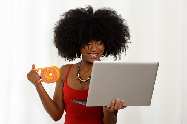 Foto mujer joven sosteniendo una computadora portátil con una taza en forma de rosquilla mirando la computadora portable adolescente afrobrasileño
