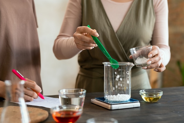 Foto mujer joven sosteniendo cristalería con masa de jabón derretido sobre la mesa con cristalería mientras hace la mezcla para productos cosméticos naturales hechos a mano