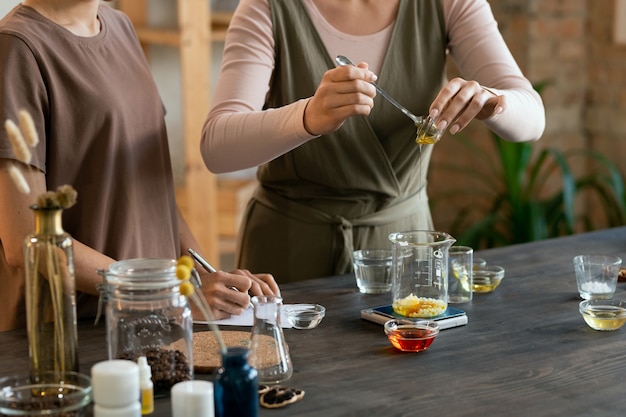 Mujer joven sosteniendo cristalería con masa de jabón derretido sobre la mesa con cristalería mientras hace la mezcla para productos cosméticos naturales hechos a mano