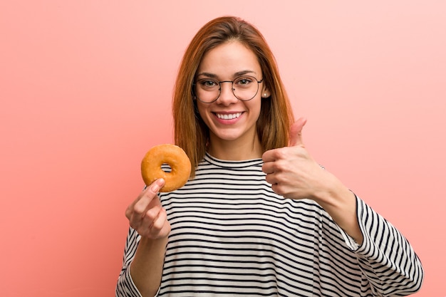 Mujer joven sosteniendo un donut sonriendo y levantando el pulgar