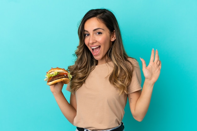 Mujer joven sosteniendo una hamburguesa sobre antecedentes aislados saludando con la mano con expresión feliz